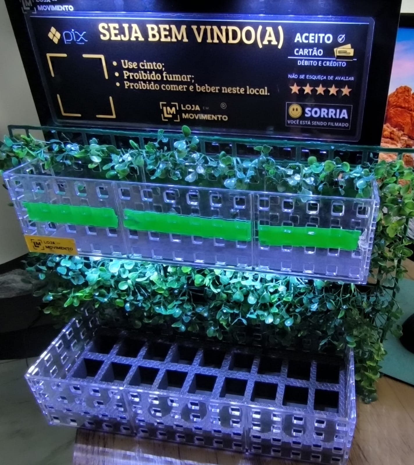 Bandeja de perfumes com placa de boas-vindas e parede de grama sintética iluminada por LED, sobre bancada, mostrando o acabamento do kit fora do carro.